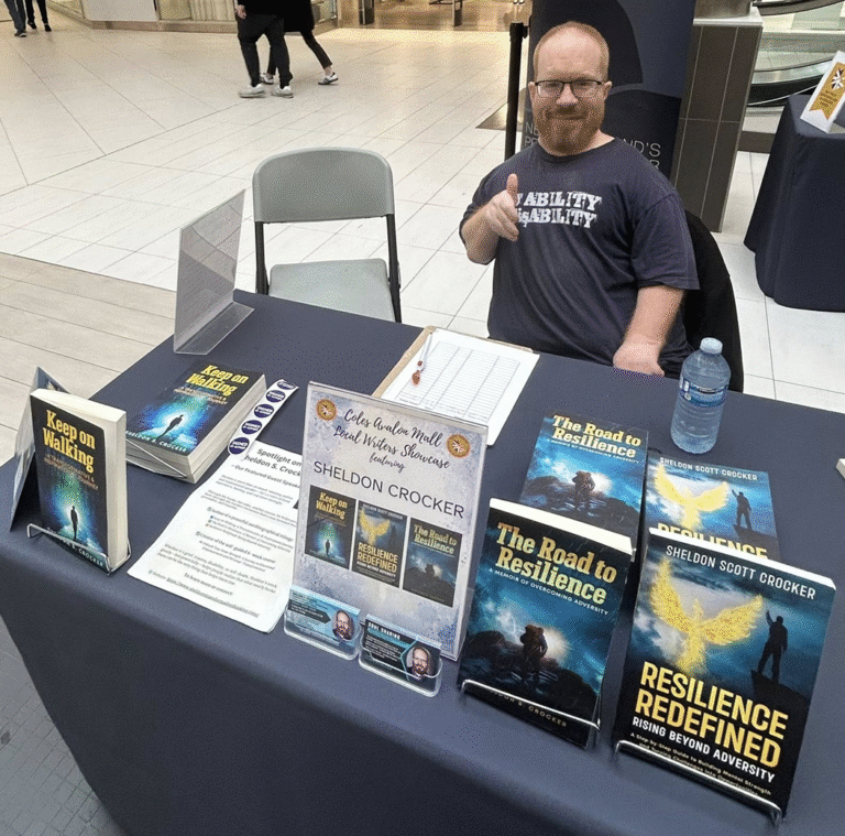 Sheldon S. Crocker sitting at his book signing table in a shopping mall, smiling and giving a thumbs-up beside his three books — Keep on Walking, The Road to Resilience, and Resilience Redefined — displayed neatly on the table with promotional materials.